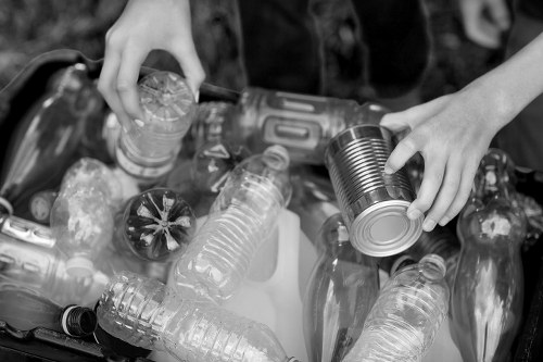 A close-up of two hands collecting recyclable waste, with one hand holding a silver metal tin can and the other grasping a clear plastic bottle, as part of a waste segregation activity outdoors. The background shows additional plastic bottles and containers in a pile, indicating a recycling or rubbish collection effort likely associated with waste management services such as those offered by Office Clearance Wembley in the Wembley area. The objects are situated on a surface, possibly a worktable or the ground, with natural lighting highlighting the different textures of the metal and plastic materials. The image emphasizes the importance of sorting recyclable rubbish, aligning with rubbish removal and recycling services available in the Wembley region to maintain cleanliness and environmental responsibility.