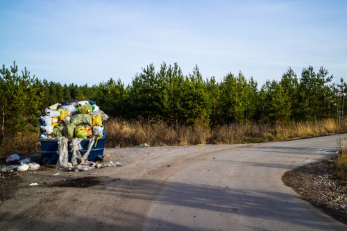 A blue waste collection bin situated on a corner of a rural dirt road surrounded by scattered rubbish, including plastic bags and other refuse, with a backdrop of dense green trees and a clear, lightly clouded sky. The bin is overflowing with black and yellow plastic bags, cardboard, and various discarded items, some of which have spilled onto the ground. The scene indicates improper waste disposal in an outdoor environment near natural foliage, emphasizing the importance of professional rubbish removal services consistent with local requirements in the Wembley area. The dirt road curves gently to the right, with visible tire tracks, and the overall atmosphere suggests a quiet, undeveloped area, highlighting the need for environmentally responsible waste management by companies like Office Clearance Wembley to keep outdoor spaces tidy and compliant with regulations.