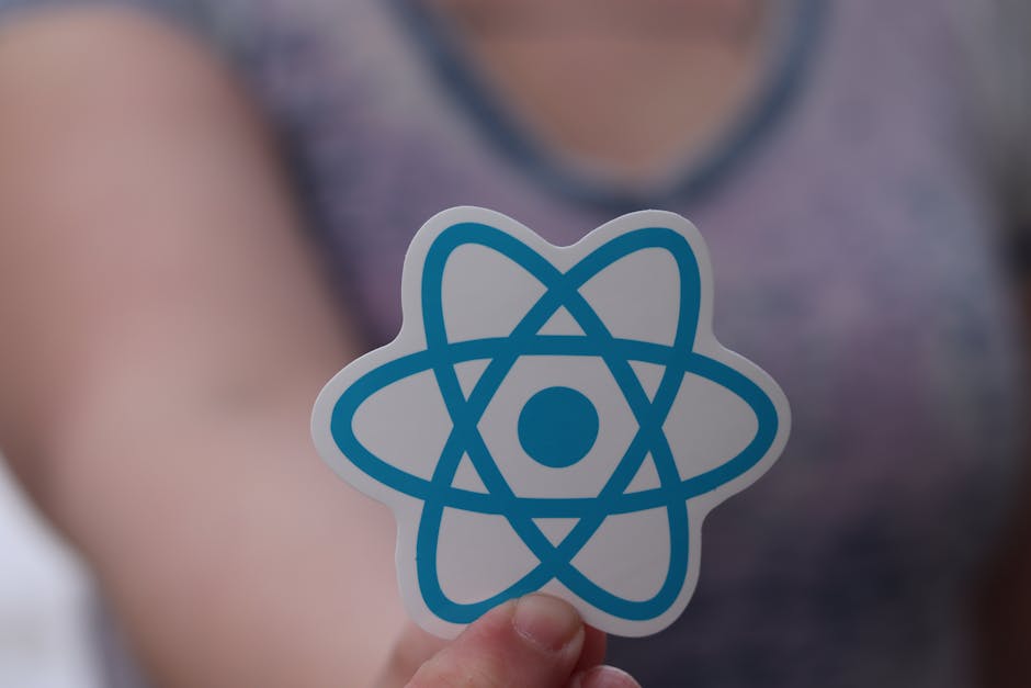 A person holding a small, circular sticker featuring a blue cartoonish atomic symbol with a central dot and three orbiting loops. The individual’s fingers are visible grasping the sticker, with a bl